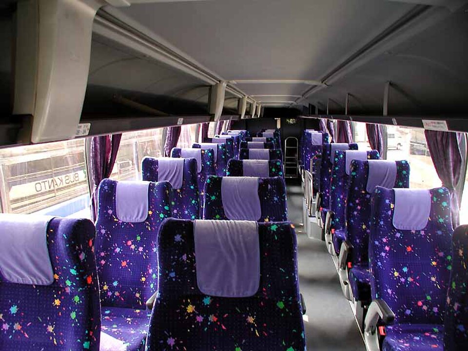 Inside a highway bus: rows of reclining seats, two on each side of the aisle, with headrest covers, window curtains and overhead luggage racks